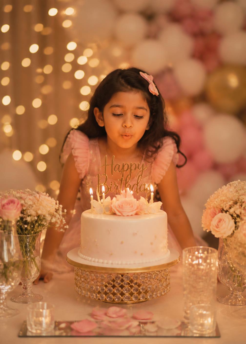Child blowing out candles on a pastel birthday cake surrounded by warm fairy lights