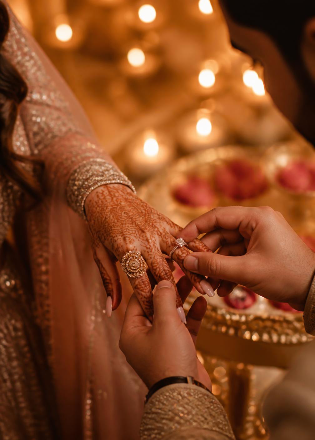 Ring exchange close-up with mehendi hands and warm candlelight during an engagement
